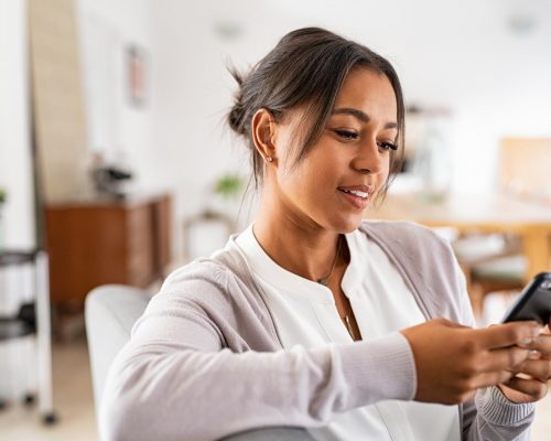 Mature african woman sitting on couch at home using smartphone. Black woman relaxing on the sofa while messaging with a cellphone at home. Happy mature indian woman typing on mobile phone in living room.